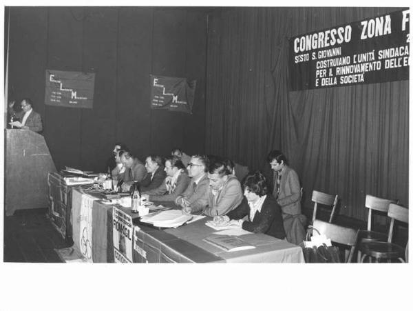 Congresso Fiom Cgil zona Sesto San Giovanni - Interno - Palco - Tavolo della presidenza con, tra gli oratori, Antonio Pizzinato - Giovanni Chinosi al microfono - Parola d'ordine - Bandiera Flm - Manifesti - Bandiera Cgil Camera del Lavoro