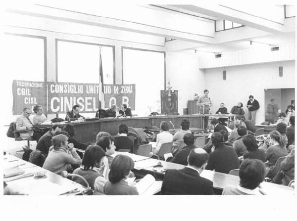 Assemblea costitutiva del Consiglio unitario sindacale della zona di Cinisello Balsamo - Interno - Tavolo della presidenza - Delegato al microfono - Striscione - Bandiera italiana - Platea con pubblico