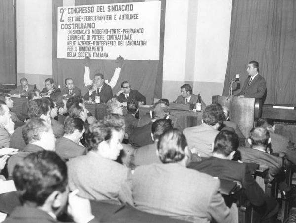 Camera del Lavoro - Sala "Bruno Buozzi" - Interno - Secondo congresso del sindacato, settore ferrotranvieri e autolinee - Tavolo della presidenza con oratore al microfono - Platea di spalle - Parola d'ordine del congresso