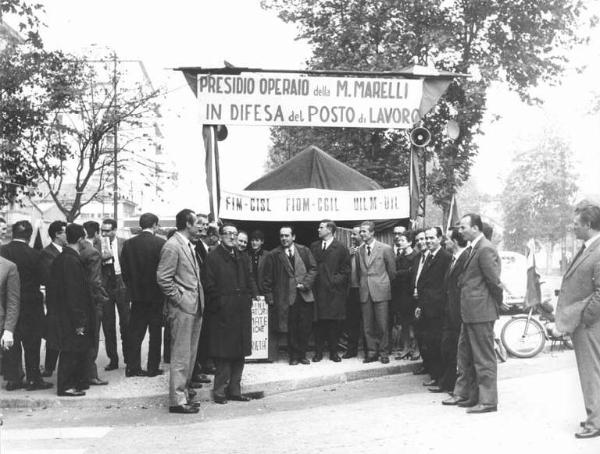 Presidio operaio alla Magneti Marelli in difesa del posto di lavoro - Visita di una delegazione sindacale - Foto di gruppo davanti alla tenda, tra i sindacalisti: L. Banfi, A. Pizzinato, P. Boni, P. Perotta, B. Trentin, A. Breschi - Striscione