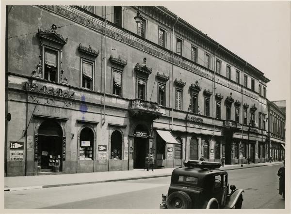 Milano - Via Verdi n.4 - Palazzo Talenti // insegne di negozi, frecce indicatrici rifugio antiaereo, automobile in primo piano, manifesti propagandistici fascismo e monarchia,due militari in uniforme visibili a distanza