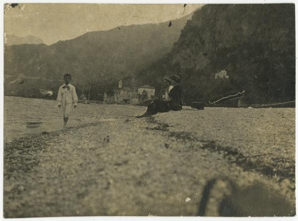 Ritratto di gruppo - Elvira Lazzaroni seduta sulla spiaggia con altra donna e bambino sulla riva - Lago, veduta - Montagne sullo sfondo