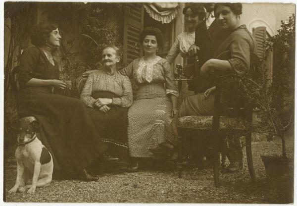 Ritratto di gruppo - Elvira Lazzaroni con altre due donne e anziana sedute, donna e uomo con macchina fotografica a soffietto in mano - Esterno, cortile - Cane