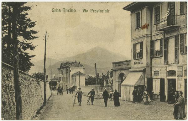 Erba - Incino - Via Provinciale - Gruppo di persone sulla strada, passanti - Edifici, bar - Veduta delle montagne sullo sfondo