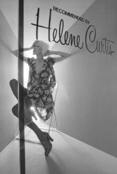 Helene Curtis. Modella posa seduta su uno sgabello indossando un abito a fiori.