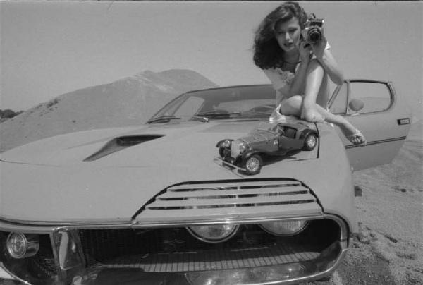 Ritratto femminile - giovane - modella che fotografa un modellino Alfa Romeo seduta sul cofano di un'automobile. Anna