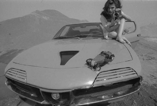 Ritratto femminile - giovane - modella che fotografa un modellino Alfa Romeo seduta sul cofano di un'automobile. Anna