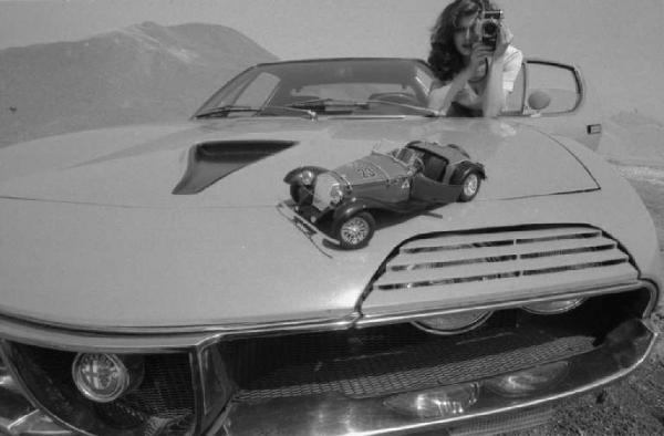 Ritratto femminile - giovane - modella che fotografa un modellino Alfa Romeo appoggiato sul cofano di un'automobile. Anna