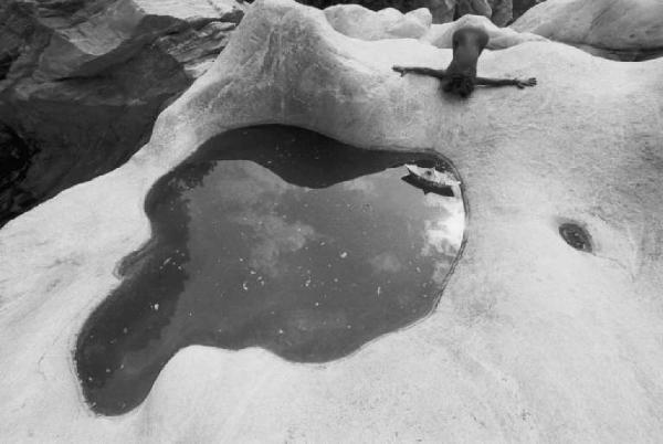 Modella nuda accanto a pozza d'acqua, sdraiata su rocce sagomate da un torrente