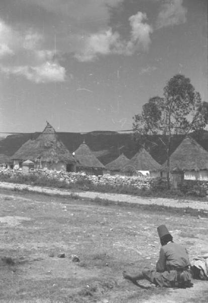 Viaggio in Africa. Villaggio - capanne con tetto di paglia - militare indigeno seduto per terra in primo piano