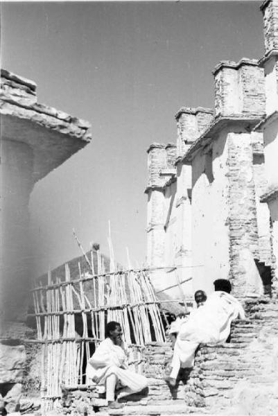 Viaggio in Africa. Tre indigeni siedono sulle gradinate in pietra che conducono ad un edificio in muratura