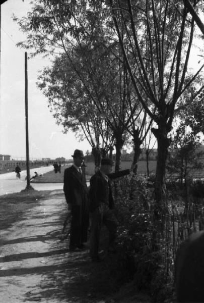 Periferia di Milano: persone che osservano il campo della bocciofila attraverso un filare di alberi