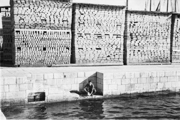 Viaggio in Jugoslavia. Gruz (Gravosa): bancali con assi accatastate lungo la banchina del porto con uomo che lava un utensile in acqua