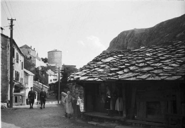 Viaggio in Jugoslavia. Mostar: scene di vita quotidiana - coppia di militari percorre una strada mentre sulla soglia di un'abitazione vi sono alcune donne