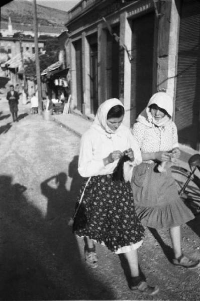 Viaggio in Jugoslavia. Mostar: scene di vita quotidiana - coppia di donne lavora all'uncinetto mentre percorre una strada del paese
