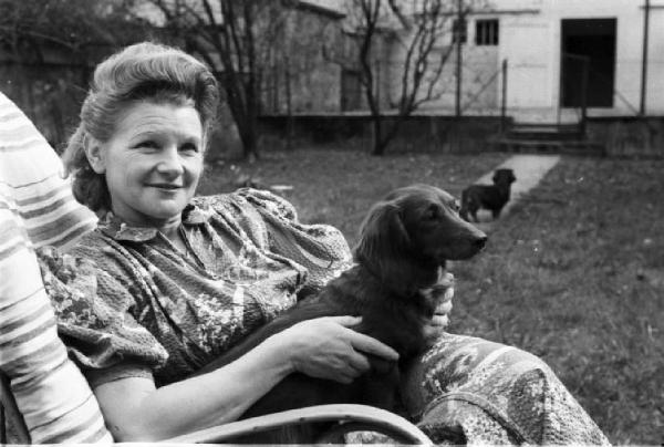 Internamento in Svizzera. Attisholz. Ritratto femminile, Milly siede in giardino con in braccio due cani bassotto. Indossa un vistoso abito fantasia