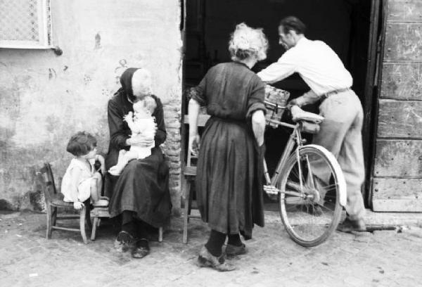 Roma - isola Tiberina. Nucleo famigliare nei pressi dell'entrata di casa: una anziana donna tiene in braccio un bimbo neonato mentre al suo fianco un giovane fanciullo osserva la madre e il nonno entrare in casa con la bicicletta