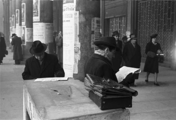 Italia Dopoguerra. Milano - piazza Vetra - Scorcio nei pressi di un edificio porticato sulla piazza - in primo piano una coppia di eleganti signori mentre leggono alcuni documenti