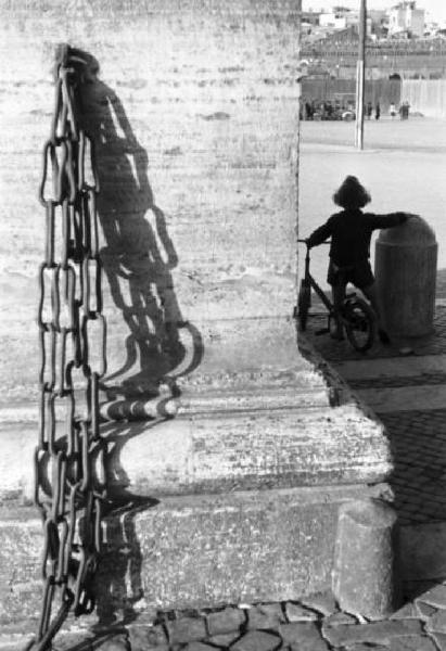Roma. Nei pressi di Piazza San Pietro una bambina in bicicletta si appoggia ad un paracarro. In primo piano scorcio di un pilastro con catena