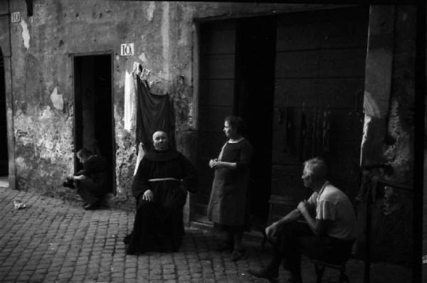 Italia Dopoguerra. Roma - Quartiere Trastevere - un frate siede sulla soglia di un'abitazione con alcune persone