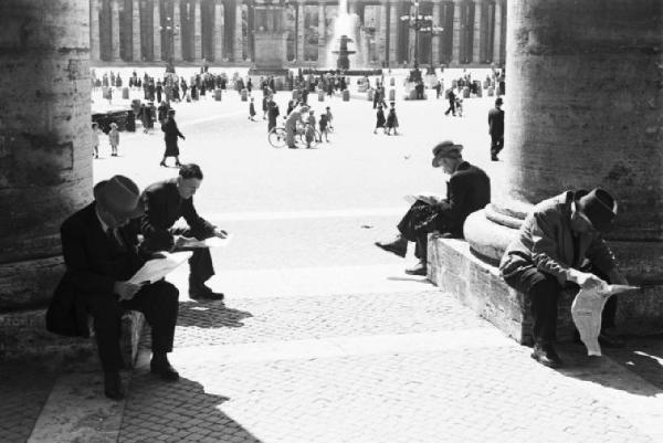 Roma. Piazza San Pietro. Alcuni uomini siedono ai piedi del colonnato leggendo il quotidiano; sullo sfondo la piazza