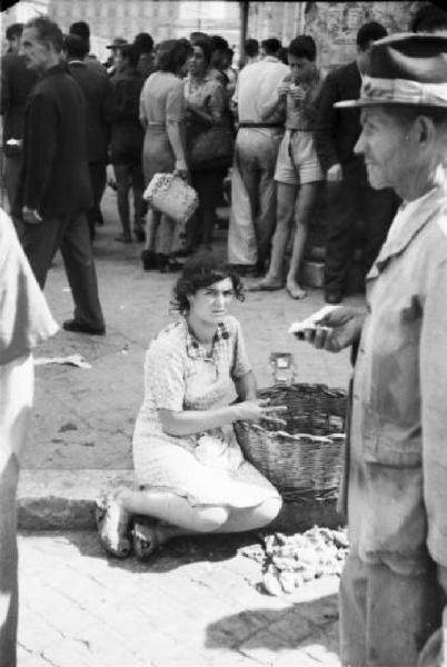 Italia Dopoguerra. Roma - Una donna siede con un cesto di prodotti alimentari in una piazza - Alle sue spalle un gruppo di persone