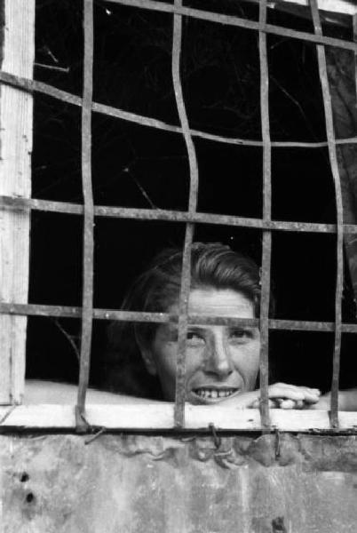 Italia Dopoguerra. Milano - Periferia - Baraccopoli - Ritratto femminile, donna che sorride attraverso le grate a protezione della finestra di una baracca