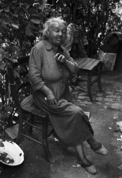 Italia Dopoguerra. Milano - Periferia - Baraccopoli - Ritratto femminile, donna che posa accanto a un cagnolino accucciato su una lapide utilizzata come tavolo