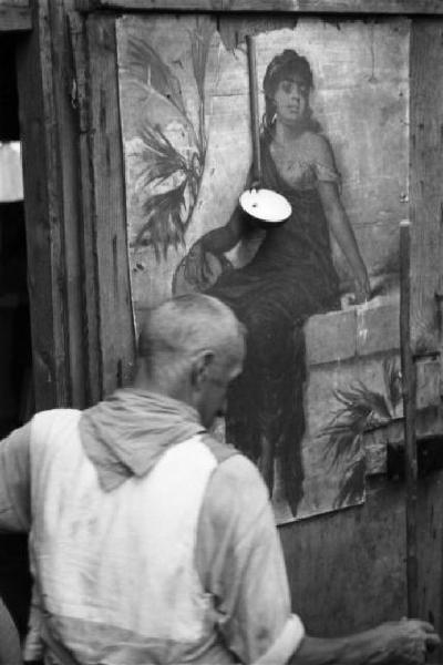 Italia Dopoguerra. Milano - Periferia - Baraccopoli - Uomo anziano di spalle e la parete esterna di una baracca dipinta con una figura femminile