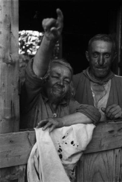 Italia Dopoguerra. Milano - Periferia - Baraccopoli - Ritratto di coppia, donna anziana deforme che indica affacciata alla finestra di una baracca accanto a un uomo anziano