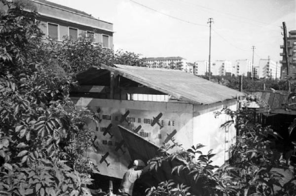 Italia Dopoguerra. Milano - Periferia - Scorcio di una baraccopoli - Baracca costruita con materiali di recupero tra cui un pannello di propaganda militare