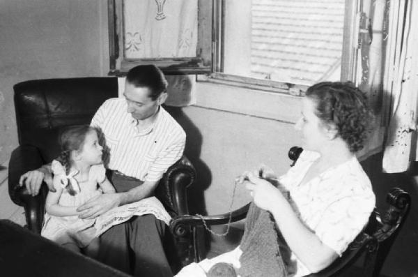 Italia Dopoguerra. Milano - Scene di vita famigliare - un uomo siede in poltrona con la figlia mentre la moglie si dedica al lavoro a maglia