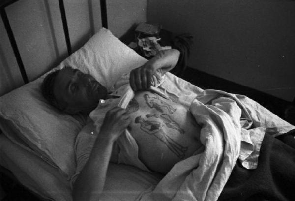 Italia Dopoguerra. Milano - Albergo popolare Camera da letto, interno - Ospite a letto mostra il petto tatuato