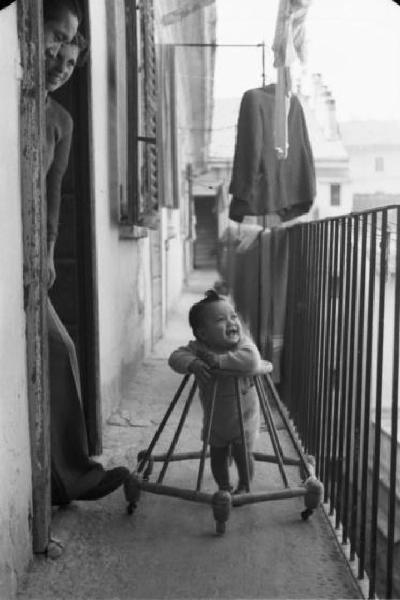Italia Dopoguerra. Milano - Quartiere cinese - bambino cinese si muove con il girello sul ballatoio di una casa a ringhiera sotto lo sguardo attento di una coppia di persone