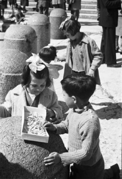 Roma. Piazza San Pietro. Coppia di bambini osserva il contenuto di una scatola appoggiata a un panettone in pietra