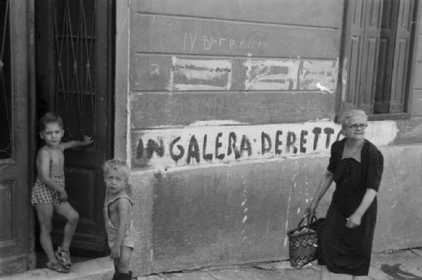 Italia Dopoguerra. Trieste - Anziana donna cammina rasente un muro riportante la scritta "IN GALERA DERETT - - -" (sic) - Accanto un bambino sulla soglia di casa