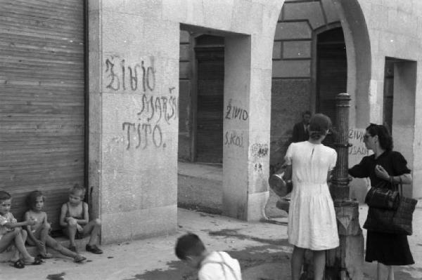 Italia Dopoguerra. Trieste - Donne che conversano e ragazzini che giocano nei pressi di un muro riportante scritte inneggianti a Tito [?]