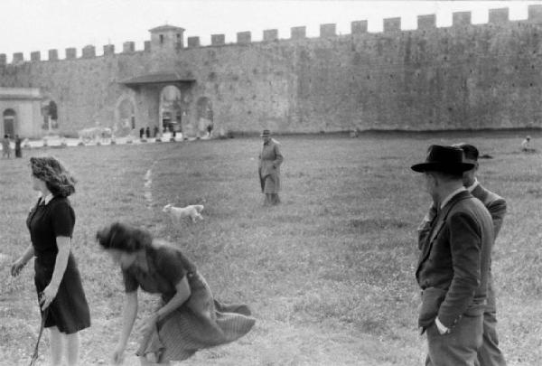 Pisa. Giovani donne uomini e cagnolini passeggiano sul prato - sullo sfondo le mura antiche