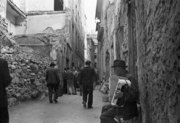 Italia Dopoguerra. Genova - Scorcio di un carrugio con edifici semidistrutti dai bombardamenti - in primo piano un uomo anziano suona una fisarmonica