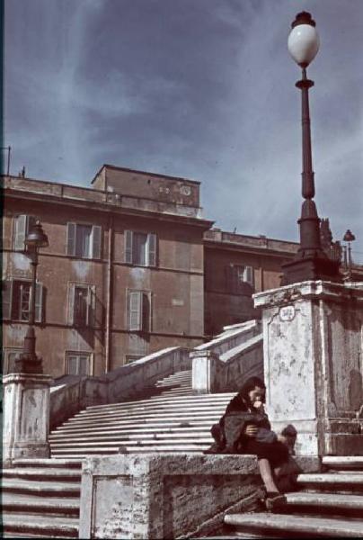Roma. Donna con bambino in braccio siede lungo la scalinata di Trinità dei Monti in Piazza di Spagna. Sullo sfondo alcuni edifici con intonaco rosso - "case rosse"