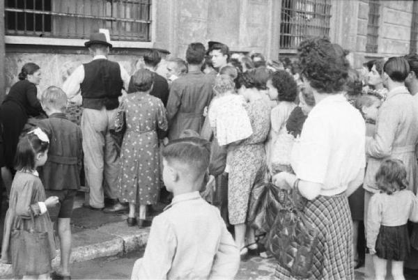 Italia Dopoguerra. Milano. Vendita al mercato di prodotti a prezzi calmierati. Folla di ripressa di spalle davanti a un edificio