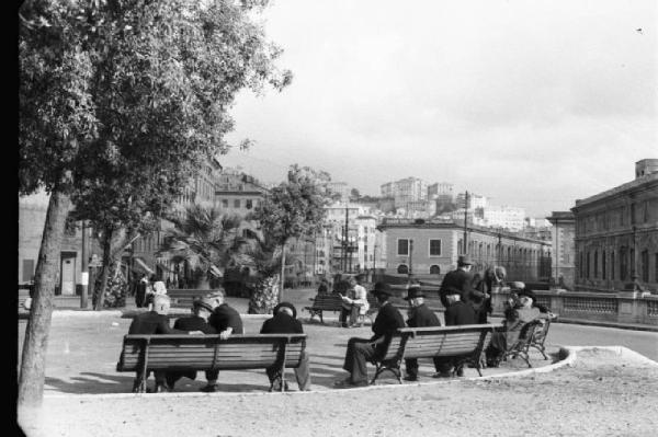 Italia Dopoguerra. Genova - Scorcio di una piazza con gruppo di uomini sulle panchine dei giardini pubblici
