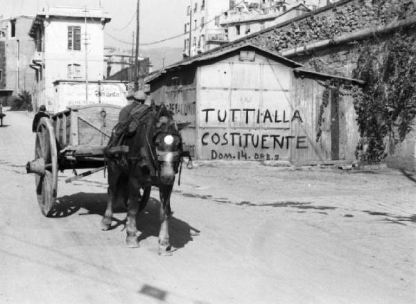 Italia Dopoguerra. Genova - Scorcio di una strada con un carro trainato da un cavallo - Sulla facciata di una casa si nota la scritta "tutti alla costituente"