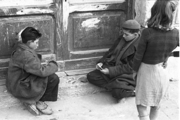 Italia Dopoguerra. Terracina. Due ragazzini giocano a carte sulla soglia di un portone. Una bambina li osserva