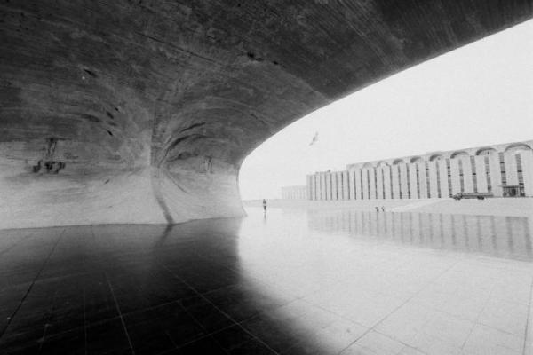 Brasilia. Spianata lastricata con palazzo sullo sfondo e coperura a vela in calcestruzzo in primo piano