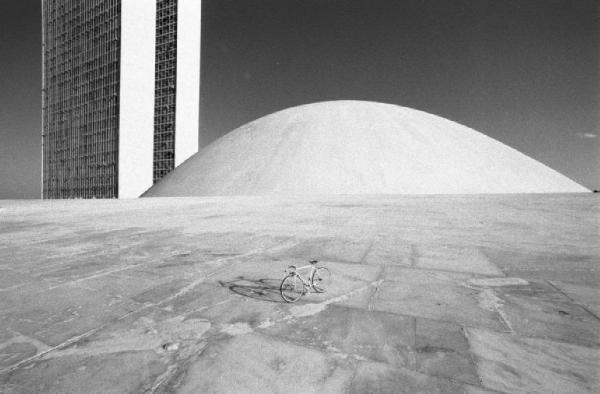 Brasilia. Bicicletta da corsa davanti a una delle due cupole del Congresso Nazionale, con i due grattacieli sullo sfondo. Si nota sul selciato l'ombra di una persona