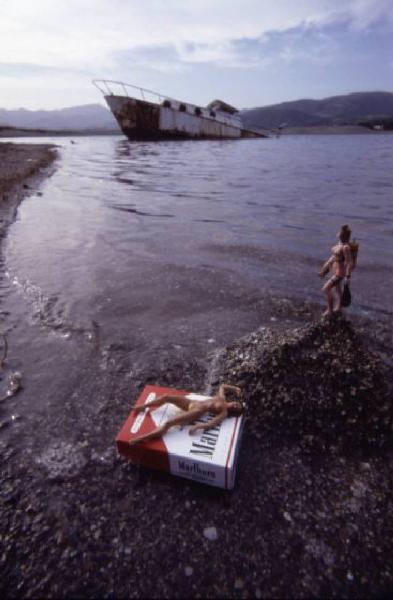 Marlboro. Pacchetto di sigarette sul bagnasciuga, con piccole bambole nude intorno e relitto di nave sullo sfondo