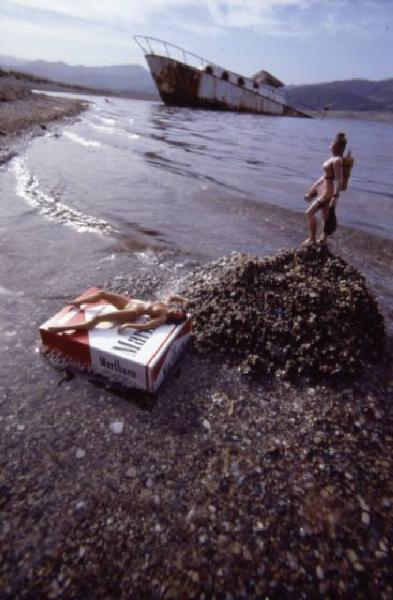 Marlboro. Pacchetto di sigarette sul bagnasciuga, con piccole bambole nude intorno e relitto di nave sullo sfondo