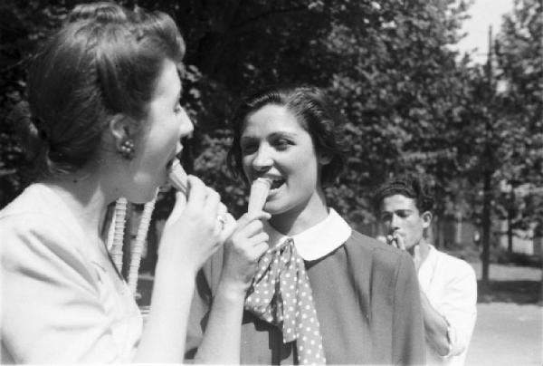 Milano. Parco Sempione. Due giovani donne mangiano il gelato scherzando - alle loro spalle il gelataio fuma una sigaretta
