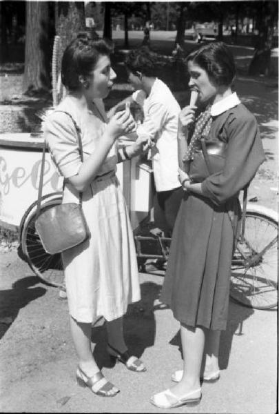 Milano. Parco Sempione. Due giovani donne mangiano il gelato chiacchierando - alle loro spalle il carretto del gelataio